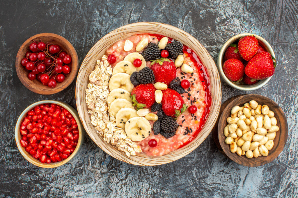 Loaded oatmeal bowl topped with sliced bananas, fresh strawberries, blackberries, peanuts, and rolled oats, surrounded by small bowls of pomegranate seeds and berries.